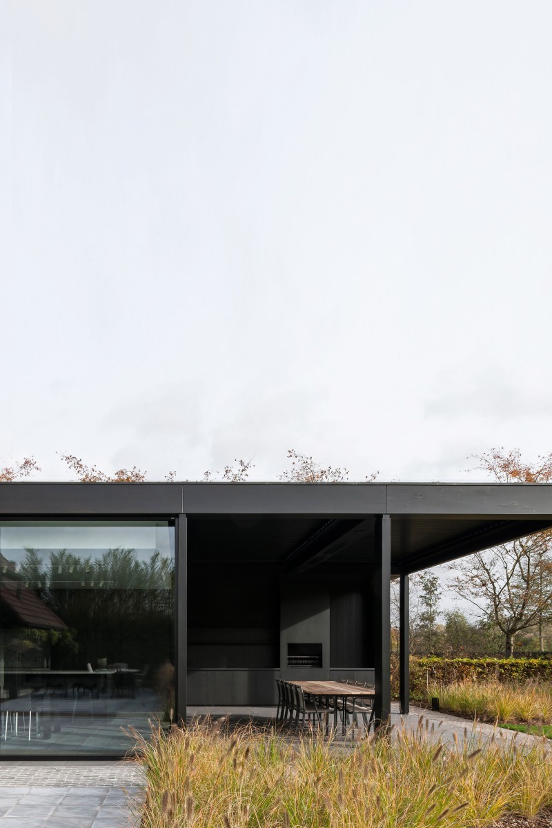 Une maison pavillon minimaliste foncée aux tons monochromes avec toit plat et terrasse couverte implantée dans un pré de graminées.